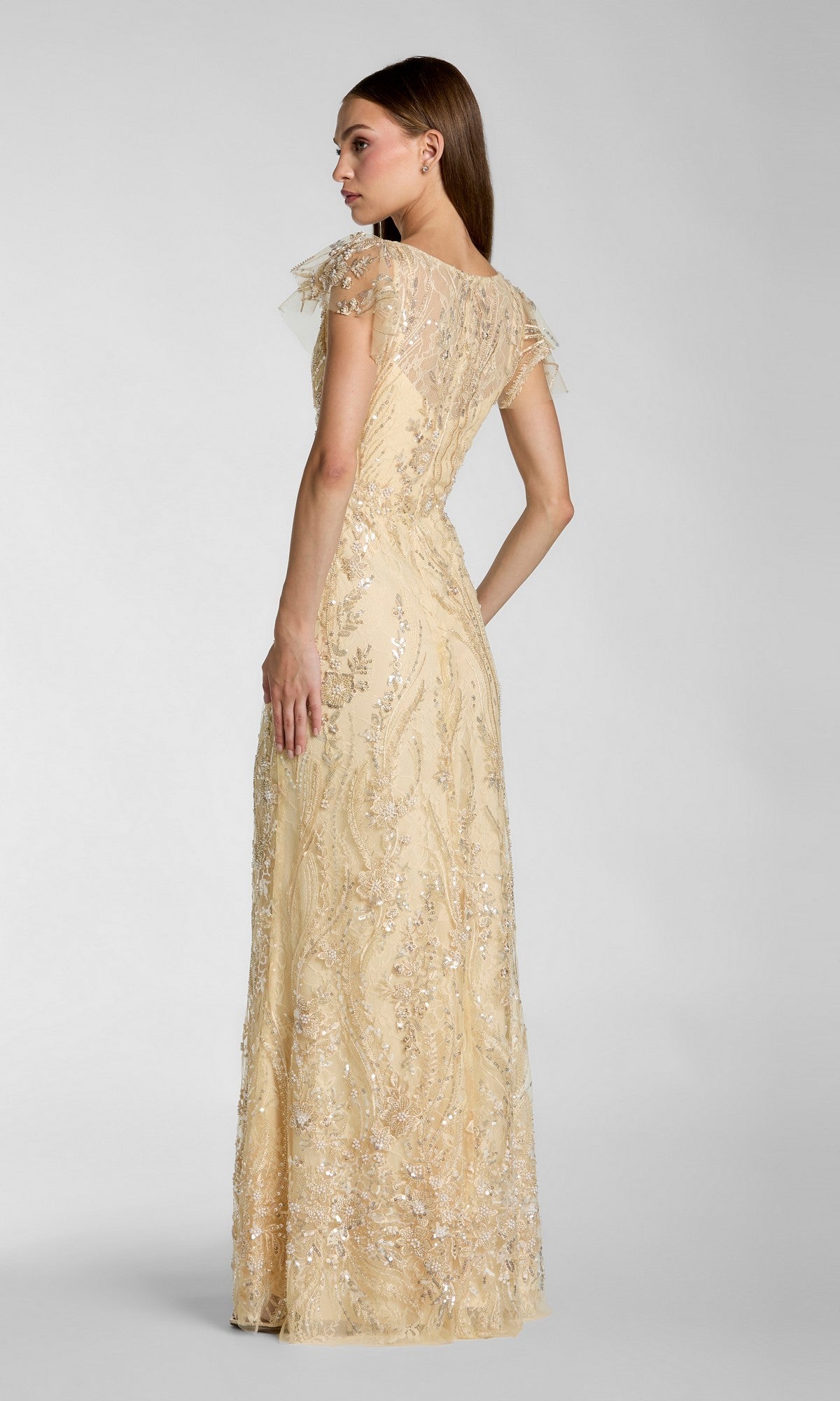 A woman stands facing away from the camera against a plain gray background, wearing the Long Formal Dress: Mac Duggal 68914—a floor-length, short-sleeved beige lace gown adorned with sequin detailing that reflects Mac Duggal’s signature elegant style.